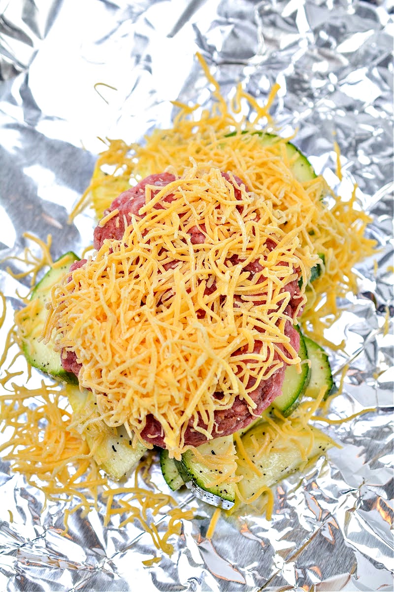 Shredded cheese on top of raw burger and veggies in a foil packet